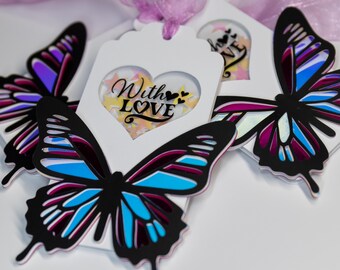 May include: Three white gift tags with a butterfly design. The center tag features a heart-shaped window with glitter and the text "With LOVE".