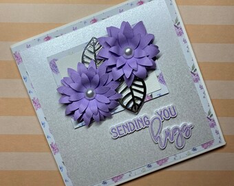 May include: A handmade greeting card with a silver glitter background and a purple floral design. The card says "Sending you hugs".