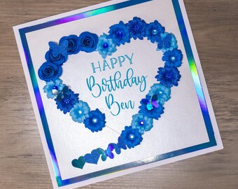 May include: A white card with a blue and turquoise paper flower heart design. The card says "Happy Birthday Ben" in a white script font.