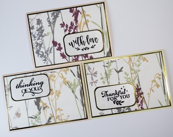 May include: Three greeting cards with floral patterns in muted colors of gray, purple, and gold. Each card has a white label with a different message in black script: "with love," "thinking of you," and "thankful for you."