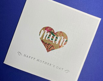 May include: A white card with a heart-shaped cutout filled with patterned paper. The word "mum" is written in white letters on the patterned paper. The card reads "Happy Mother's Day" at the bottom.
