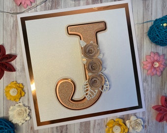 May include: A white card with a copper border, featuring a large, glittery, copper-colored letter "J" decorated with three brown paper roses and leaves.