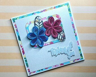May include: A white card with a blue and pink paper flower design. The card has a blue and white patterned border and the text "Thinking of you" in white.