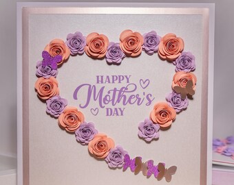 May include: A handmade card with a heart-shaped arrangement of peach and purple paper flowers. The card has a white background with a silver border and the text "Happy Mother's Day" in purple lettering.