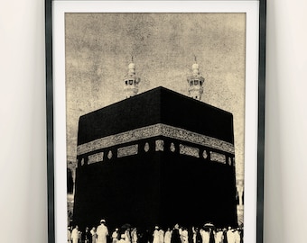 Op de afbeelding: Een ingelijste wandkunstprint met een contrastrijke, monochrome afbeelding van de Kaaba in Mekka. De afbeelding toont het grote, zwarte, kubusvormige bouwwerk met gedetailleerde goudkleurige kalligrafiebanden, omringd door mensen in witte kleding.