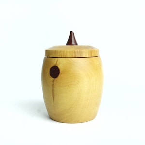 Peut inclure: Une boîte en bois avec un couvercle et un petit bouton en bois sur le dessus. La boîte est de couleur brun clair avec un cercle brun foncé sur le côté.