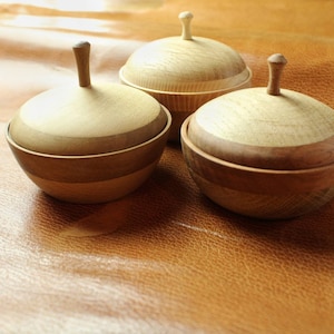 Peut inclure: Trois bols en bois avec couvercles, chacun avec un motif de grain de bois différent. Les bols sont de couleur brun clair et ont une finition lisse et polie.