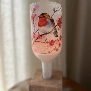 Könnte beinhalten: Eine weiße Glasflasche mit einem Design aus Rotkehlchen und roten Beeren, die auf einem Holzsockel steht. Die Flasche wird von innen beleuchtet und erzeugt ein warmes Leuchten.