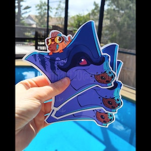 May include: Three blue and purple stickers featuring a cartoon manta ray with a smaller cartoon fish on its back. The manta ray has a cookie in its mouth and the stickers are stacked on top of each other.