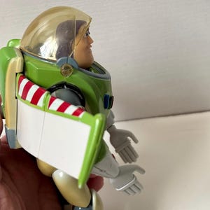 以下が含まれることがあります： 緑と白の宇宙服と透明なヘルメットを身に着けたバズ・ライトイヤーのアクションフィギュア。フィギュアは笑顔で腕を広げ、翼には赤と白のストライプのアクセントが付いています。