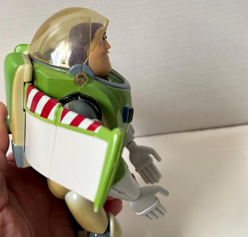 以下が含まれることがあります： 緑と白の宇宙服と透明なヘルメットを身に着けたバズ・ライトイヤーのアクションフィギュア。フィギュアは笑顔で腕を広げ、翼には赤と白のストライプのアクセントが付いています。