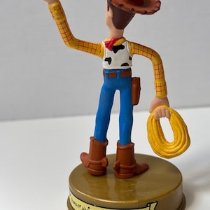 May include: A Woody figurine from the Toy Story franchise. The figure is wearing a brown cowboy hat, yellow plaid shirt, cow-print vest, blue jeans, and brown boots. The figure is holding a yellow rope and stands on a gold base with the text "Walt Disney World".
