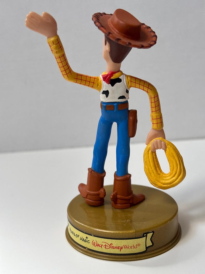 May include: A Woody figurine from the Toy Story franchise. The figure is wearing a brown cowboy hat, yellow plaid shirt, cow-print vest, blue jeans, and brown boots. The figure is holding a yellow rope and stands on a gold base with the text "Walt Disney World".