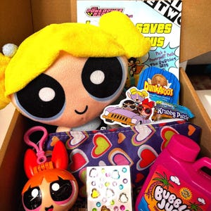 May include: A Powerpuff Girls themed gift box filled with merchandise. Includes a plush Buttercup, a Blossom figurine, a purple heart-patterned pouch, stickers, and a pink Bubble Jug. A comic book and snacks are also visible.