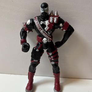 May include: A black action figure with red accents, wearing a mask with white eye markings. The figure features a red belt with a white skull buckle, red boots, silver spikes on the left shoulder, and a silver ammunition belt across the chest.