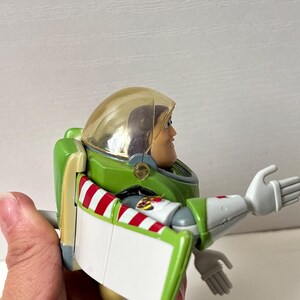 以下が含まれることがあります： 緑と白の宇宙服に赤と白のストライプのアクセントが付いた、バズ・ライトイヤーのアクションフィギュア。 透明なヘルメットと片腕を伸ばしたポーズをしています。