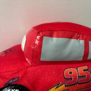 May include: A red plush toy car featuring the number 95 in orange and yellow on the side. The toy has smooth, shiny material for the windows and windscreen, with a soft, fuzzy red fabric body. Black tyres with white text are visible at the base.
