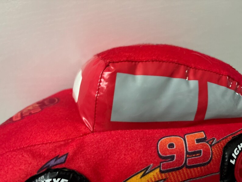 May include: A red plush toy car featuring the number 95 in orange and yellow on the side. The toy has smooth, shiny material for the windows and windscreen, with a soft, fuzzy red fabric body. Black tyres with white text are visible at the base.