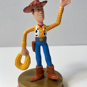May include: A Woody action figure from the Toy Story franchise. The figure is wearing a brown cowboy hat, yellow plaid shirt, blue jeans, and brown boots. It is holding a yellow rope and standing on a gold-colored base with the text "Woody 1995".
