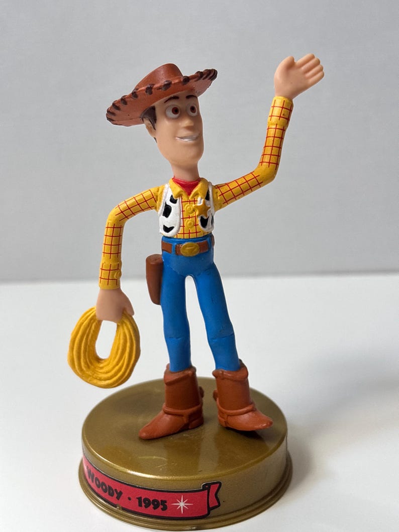 May include: A Woody action figure from the Toy Story franchise. The figure is wearing a brown cowboy hat, yellow plaid shirt, blue jeans, and brown boots. It is holding a yellow rope and standing on a gold-colored base with the text "Woody 1995".