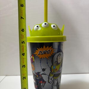 以下が含まれることがあります： トイ・ストーリーのキャラクターが描かれた透明なプラスチックタンブラー。カップは、エイリアンのデザインの緑色の蓋と黄色のストローが付いています。カップの高さは約18cmです。カップには「ZURG！」の文字がプリントされています。