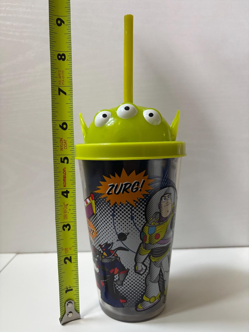 以下が含まれることがあります： トイ・ストーリーのキャラクターが描かれた透明なプラスチックタンブラー。カップは、エイリアンのデザインの緑色の蓋と黄色のストローが付いています。カップの高さは約18cmです。カップには「ZURG！」の文字がプリントされています。