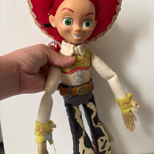 May include: A Jessie doll from the Toy Story franchise. The doll has red hair, green eyes, and a red hat. She wears a white shirt, yellow vest, and cow-print pants. The doll is approximately 12 inches tall.