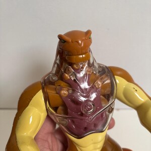 May include: A brown and yellow action figure with a clear plastic chest. The figure has a reptilian head with green eyes and a muscular build. The chest cavity reveals a purple interior. The figure is a toy.