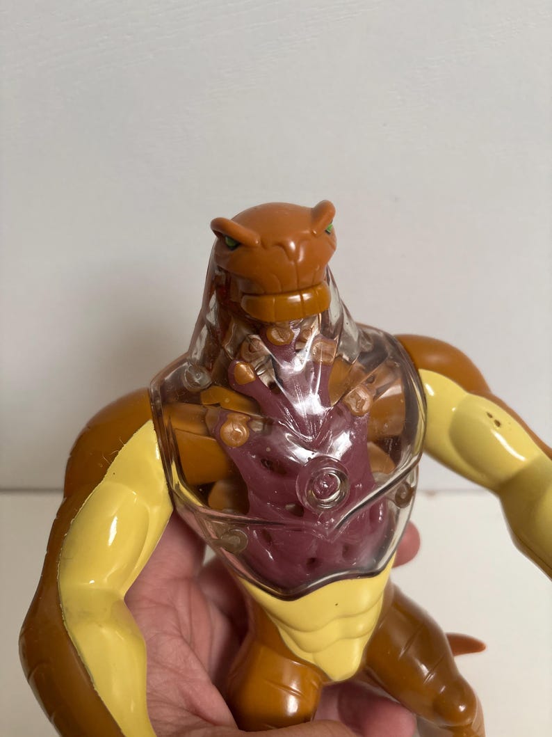 May include: A brown and yellow action figure with a clear plastic chest. The figure has a reptilian head with green eyes and a muscular build. The chest cavity reveals a purple interior. The figure is a toy.