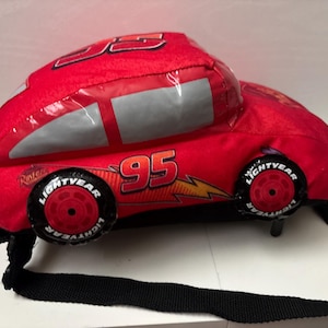 May include: A red plush backpack shaped like a race car with the number 95 printed on the side. The car features silver windows, a yellow lightning bolt graphic, and black wheels with white Lightyear text. It includes black adjustable straps.