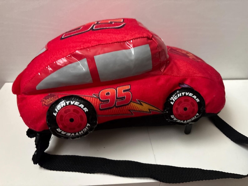 May include: A red plush backpack shaped like a race car with the number 95 printed on the side. The car features silver windows, a yellow lightning bolt graphic, and black wheels with white Lightyear text. It includes black adjustable straps.