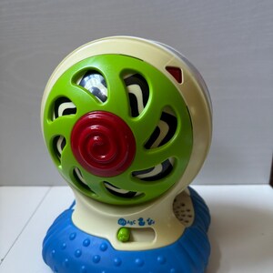 May include: A round, cream-colored plastic toy with a rotating green fan-like front featuring a red spiral center. The base is textured blue plastic. A small switch on the front has icons for Off, ABC, a duck, and music notes, with a green slider button.