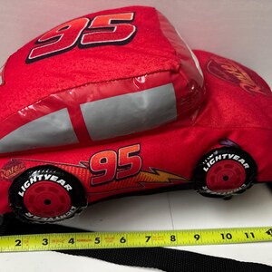 May include: A red plush car-shaped bag featuring the number 95, Rust-eze logos, and Lightyear tyres. A yellow measuring tape placed along the bottom shows the bag is approximately 33 cm long. The bag includes a black strap for carrying.