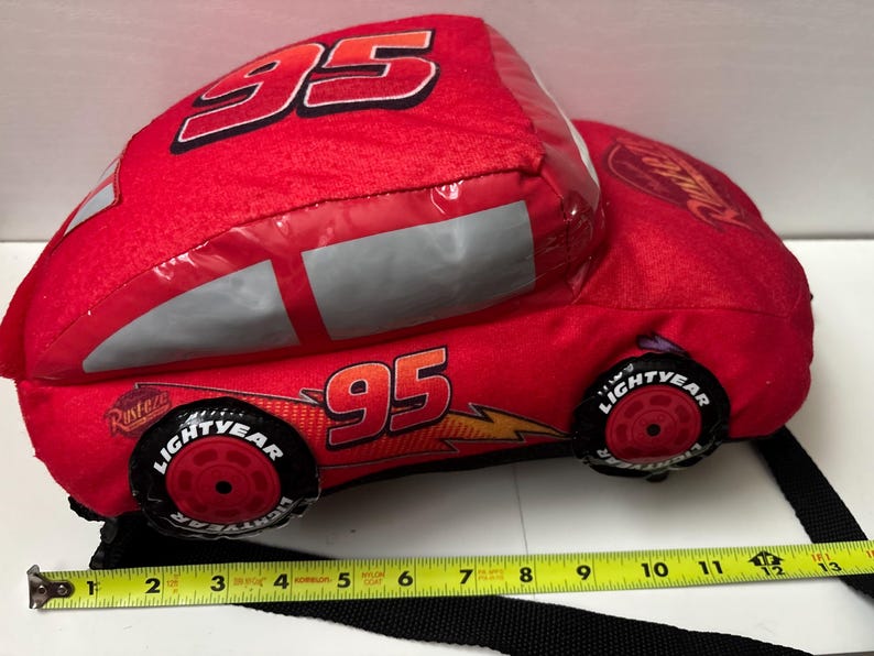 May include: A red plush car-shaped bag featuring the number 95, Rust-eze logos, and Lightyear tyres. A yellow measuring tape placed along the bottom shows the bag is approximately 33 cm long. The bag includes a black strap for carrying.