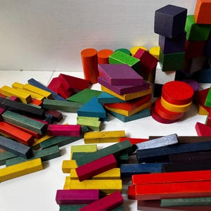 May include: A collection of colorful wooden blocks in various shapes, including cubes, rectangles, cylinders, and triangles. The blocks are painted in shades of red, yellow, green, blue, purple, and orange. The blocks are scattered on a white surface.