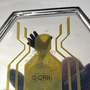 Puede incluir: Objeto transparente de forma hexagonal con un dise&ntilde;o de telara&ntilde;a amarilla. Los textos "Disney", "MARVEL", "Vietnam" y "&copy; CPII" son visibles. Probablemente un art&iacute;culo de colecci&oacute;n o promocional.