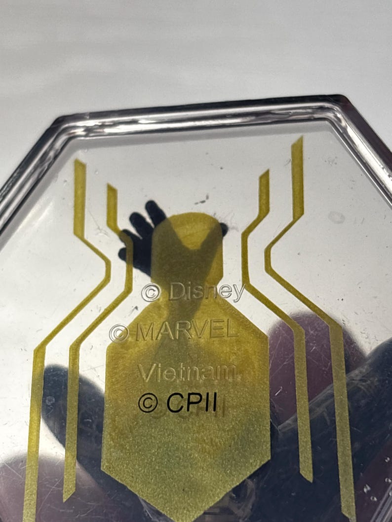 Puede incluir: Objeto transparente de forma hexagonal con un dise&ntilde;o de telara&ntilde;a amarilla. Los textos "Disney", "MARVEL", "Vietnam" y "&copy; CPII" son visibles. Probablemente un art&iacute;culo de colecci&oacute;n o promocional.