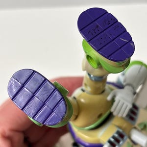 以下が含まれることがあります： バズ・ライトイヤーのアクションフィギュアのクローズアップ。フィギュアは主に黄色、緑、白で、足の裏は紫です。靴底はテクスチャード加工が施され、文字が書かれています。