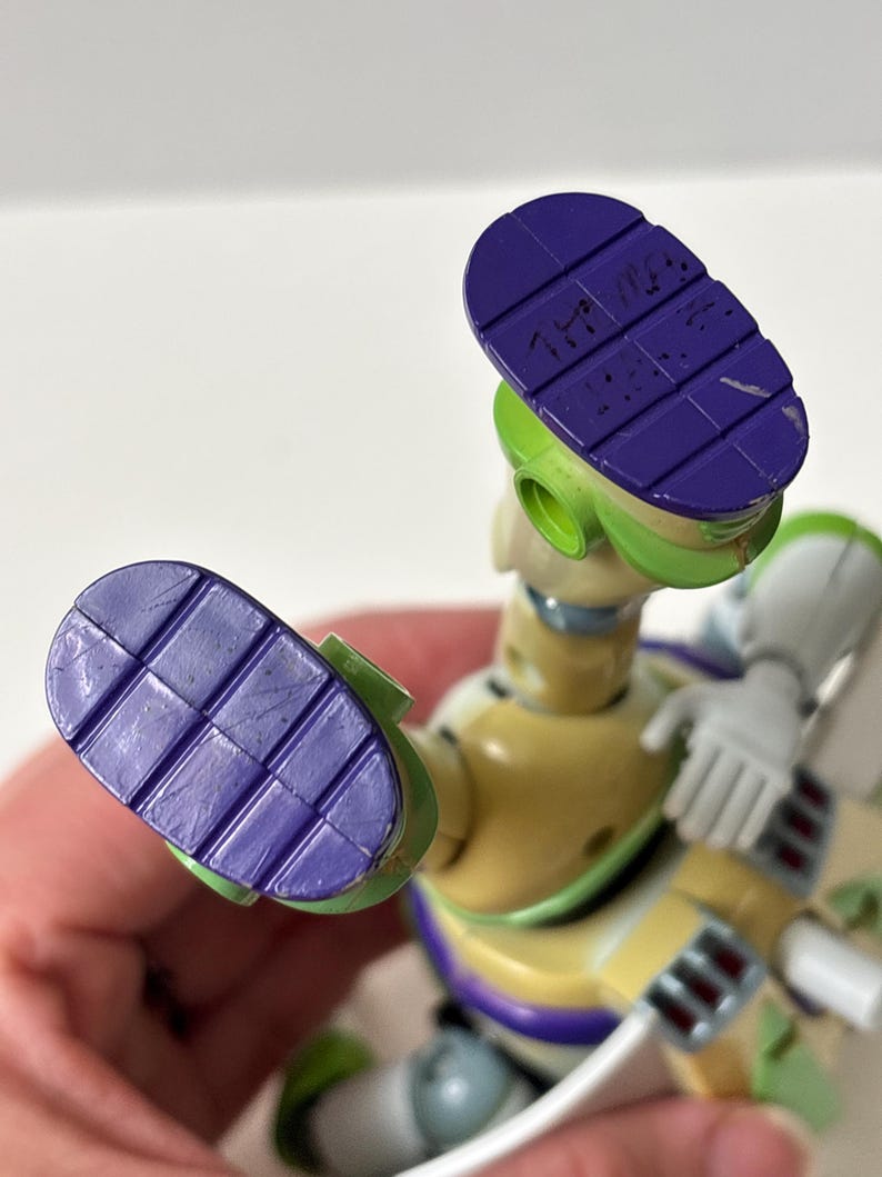 以下が含まれることがあります： バズ・ライトイヤーのアクションフィギュアのクローズアップ。フィギュアは主に黄色、緑、白で、足の裏は紫です。靴底はテクスチャード加工が施され、文字が書かれています。