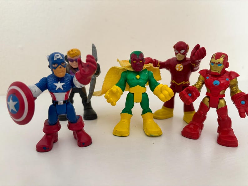 May include: A collection of colorful superhero action figures, including Captain America with his shield, Hawkeye with a bow, Vision, The Flash, and Iron Man. The figures are in various poses, showcasing their iconic costumes.