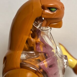 May include: An orange and clear plastic action figure of a feline-like creature. The figure has a detailed design, with visible internal structures and green eyes. The figure is posed in a dynamic stance, showcasing its articulated joints and overall design.