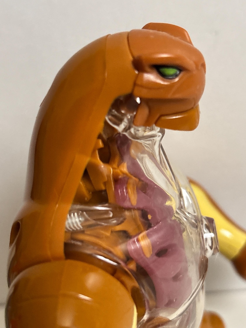 May include: An orange and clear plastic action figure of a feline-like creature. The figure has a detailed design, with visible internal structures and green eyes. The figure is posed in a dynamic stance, showcasing its articulated joints and overall design.