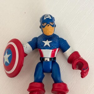 May include: A Captain America action figure with a blue and white star on the chest, red boots, and a red shield with a white star. The figure is wearing a blue helmet with a white "A" on the forehead.
