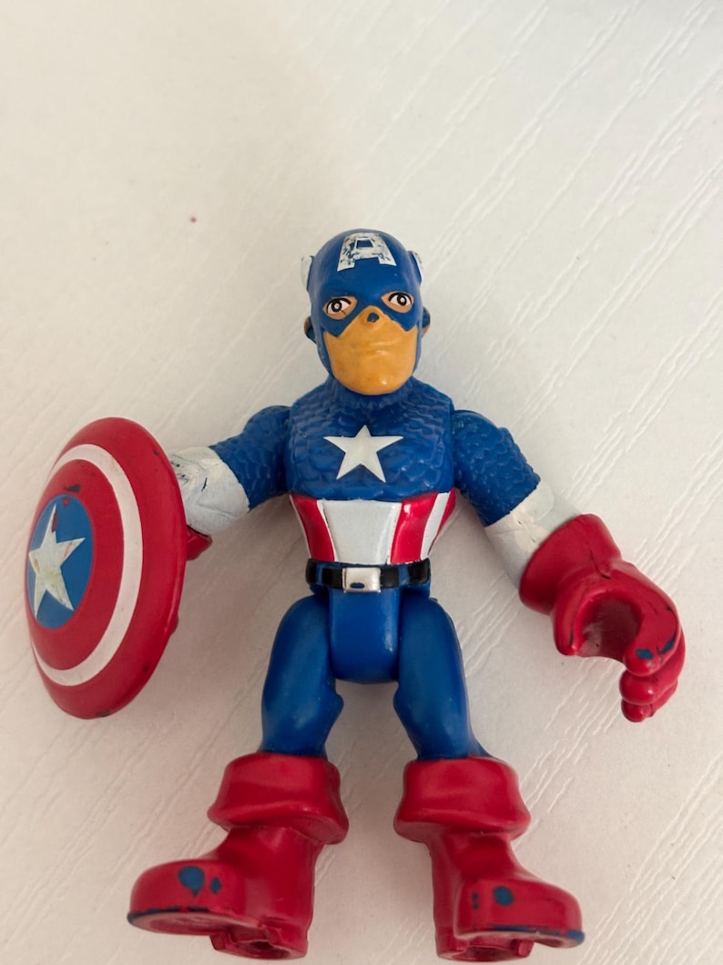 May include: A Captain America action figure with a blue and white star on the chest, red boots, and a red shield with a white star. The figure is wearing a blue helmet with a white "A" on the forehead.