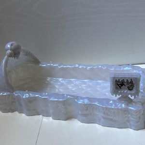 May include: A rectangular, iridescent white plastic bed frame with a textured, icy appearance. It features a rounded headboard with a skull detail and a small, rectangular television screen attached to the side, displaying a graphic of a snowboarder.