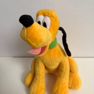 May include: A plush toy of Pluto, a yellow cartoon dog with a black nose and ears, a green collar, and a pink tongue sticking out. The toy is sitting on a white surface.