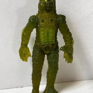 May include: A green monster action figure with a textured, scaly appearance. The figure has yellow eyes, a detailed face, and a translucent chest. The arms and legs are articulated, and the figure is standing upright.