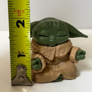 May include: A green figurine of a character from a popular science fiction franchise, wearing a tan robe with a furry collar. The figurine is approximately 2 inches tall. The character is in a meditative pose with closed eyes.