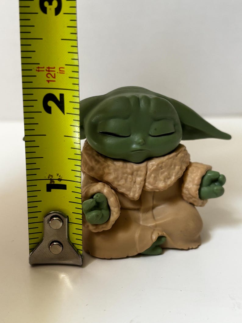May include: A green figurine of a character from a popular science fiction franchise, wearing a tan robe with a furry collar. The figurine is approximately 2 inches tall. The character is in a meditative pose with closed eyes.