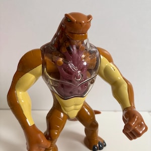 May include: A brown and yellow action figure of a reptilian humanoid alien with a clear chest and a red-purple center. The figure has a muscular build, with a tail and black claws. The figure is standing on a white surface.
